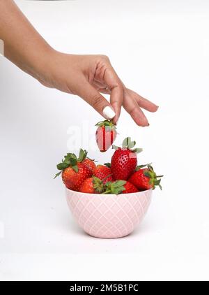 Un cliché vertical d'une main cueillant des fraises dans un bol rose sur une surface blanche Banque D'Images