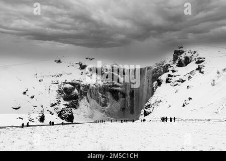 Touristes à la cascade de Skogafoss, visage de roche enneigée et glacée, nuages sombres au-dessus, photo noir et blanc, Sudurland, Islande Banque D'Images