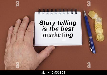 Concept d'entreprise. Sur une surface marron sont des pièces de monnaie, un stylo et un bloc-notes avec l'inscription - raconter l'histoire est le meilleur marketing. Le bloc-notes est holdin Banque D'Images
