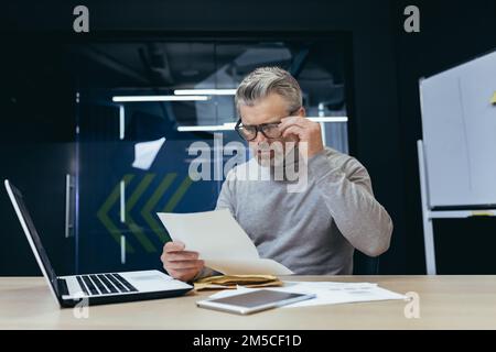 Un homme d'affaires mûr aux cheveux gris à l'intérieur d'un bureau moderne a reçu un message postal, un homme âgé contrarié ouvre une enveloppe postale, un patron lit les mauvaises nouvelles bouleversées et travaille triste au bureau avec un ordinateur portable. Banque D'Images