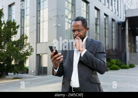 Un patron afro-américain expérimenté et expérimenté ayant lu de mauvaises nouvelles par téléphone, un homme d'affaires en costume devant un immeuble de bureaux utilisant un smartphone contrarié et déçu par la notification. Banque D'Images