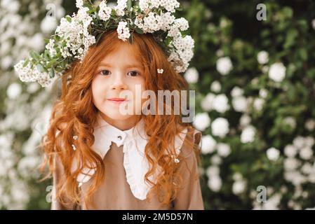 Mignon riant petite fille 3-4 ans avec de longs cheveux bouclés rouges porter une couronne florale et élégante robe rustique sur fond de nature dans outdo de jardin Banque D'Images