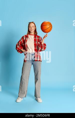 Portrait d'une jeune fille à tête rouge dans une chemise à carreaux décontractée posant, tournant le ballon de basket-ball sur le doigt sur fond bleu Banque D'Images