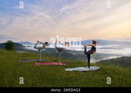 Trois femmes sportives faisant des exercices pour l'équilibre sur le tapis de yoga amonu montagnes d'été. Groupe de jeunes femmes en vêtements de sport ayant entraînement à l'extérieur ensemble. Banque D'Images