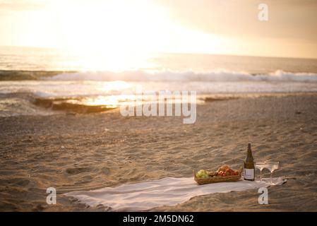 Pique-nique romantique au bord de la mer au coucher du soleil Plage déserte dans les rayons chauds du champagne du coucher du soleil et panier de fruits Banque D'Images