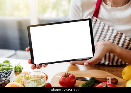 Femme mains montrant la tablette numérique blanc vide écran vide maquette à la cuisine. Chef en utilisant pc, présenter en ligne courses de nourriture sur ordinateur. Concept d'annonces d'application de livraison, livre de cuisine ou recettes de plan de régime Banque D'Images