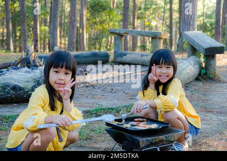 Famille heureuse, sœurs mignonnes assis sur un pique-nique près d'une tente et d'un barbecue dans une forêt de pins. Bonne famille en vacances dans la nature. Banque D'Images