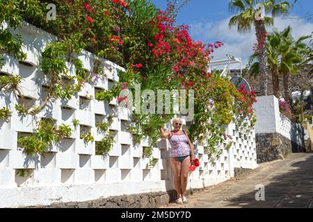 Une femme âgée se trouve près d'une clôture percée avec bougainvilliers sur la rue Magnolia à Los Gigantes, Tenerife, Canaries, Espagne. Banque D'Images