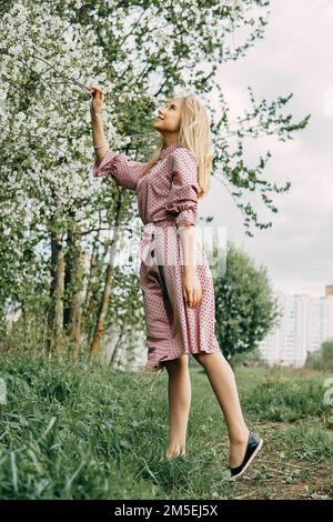 Fille blonde lors d'une promenade printanière dans le jardin avec des cerisiers en fleurs. Portrait de femme, gros plan. Une fille dans une robe à pois rose Banque D'Images