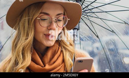 Portrait de la pensée pensive perplexe jeune fille étudiante confuse avec un téléphone dans la rue avec parapluie femme caucasienne dans le chapeau et les lunettes dans la ville Banque D'Images