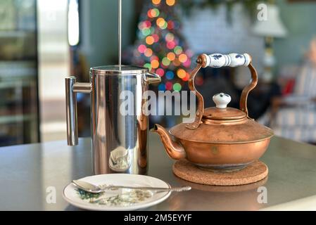 Presse française en acier inoxydable, et bouilloire à thé en cuivre sur une table en bois le matin de Noël avec de jolies lumières de Noël floues dans le Banque D'Images