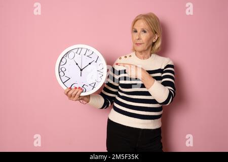 Belle femme senior tenant un réveil debout sur fond rose isolé avec le visage surprise pointant du doigt vers lui-même Banque D'Images