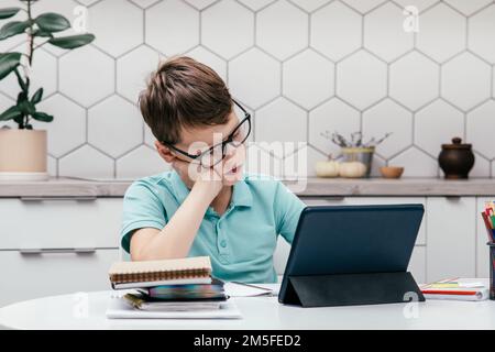 Portrait d'un jeune préadolescent concentré portant un T-shirt bleu, des lunettes, assis au bureau près des blocs-notes, livre de lecture près d'une tablette, ayant en ligne Banque D'Images