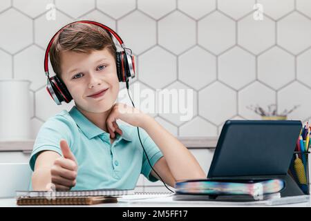 Portrait d'un jeune préadolescent souriant portant un T-shirt, un casque noir rouge, assis sur un bureau devant une tablette près d'un ordinateur portable, montrant un signe de pouce vers le haut Banque D'Images