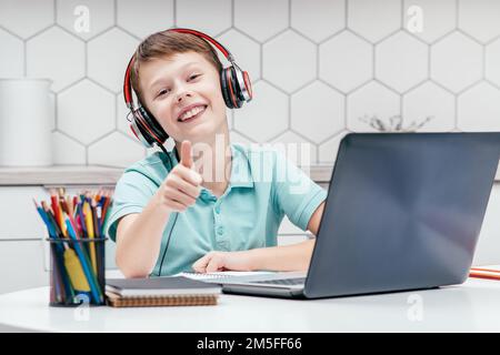 Portrait d'un jeune garçon préadolescent portant un T-shirt bleu, un casque noir rouge, assis sur un bureau devant un ordinateur portable près de la maison, montrant une affiche avec le pouce vers le haut Banque D'Images