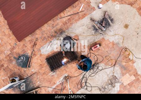 La photo aérienne de deux hommes en uniforme gris et bleu est le métal de soudage à l'arc avec machine à souder au sol sur le chantier. Banque D'Images