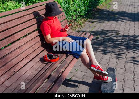 Garçon avec planche à roulettes en plastique assis sur le banc, fixe au téléphone. Banque D'Images