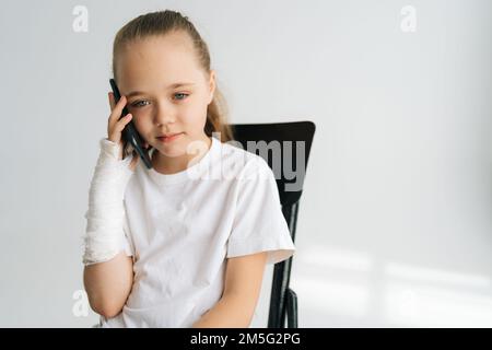 Photo moyenne d'une petite fille blonde avec un bras cassé enveloppé dans un bandage en plâtre parlant smartphone, regardant l'appareil photo, chaise assise sur fond blanc. Banque D'Images