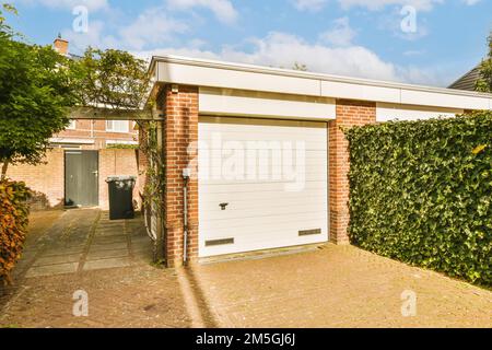 une maison en brique avec une porte de garage blanche sur le côté droit et des arbres verts dans la cour arrière derrière elle Banque D'Images