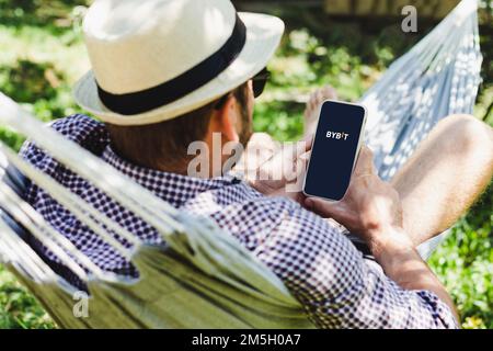 Homme dans un chapeau de paille utilisant un ordinateur portable et Bybit crypto-monnaie du marché boursier tout en se relaxant sur hamac. Banque D'Images