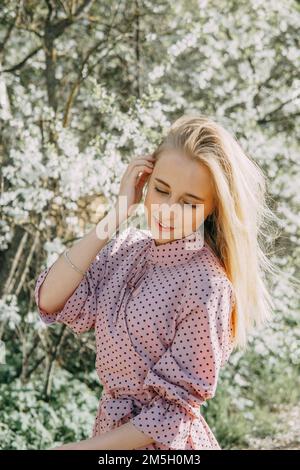 Fille blonde lors d'une promenade printanière dans le jardin avec des cerisiers en fleurs. Portrait de femme, gros plan. Une fille dans une robe à pois rose Banque D'Images