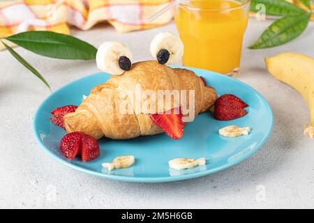 Nourriture amusante pour les enfants - croissant en forme de crabe avec fraise et banane sur une assiette bleue et un verre de jus d'orange sur fond gris Banque D'Images