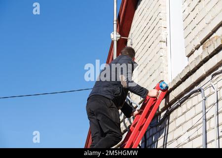 Jeune homme grimpant l'échelle. Grimpeur industriel dans le casque et travail global en hauteur. Ciel bleu. Travail risqué Banque D'Images