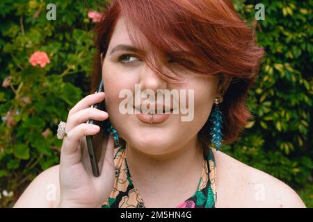 La jeune femme latina caucasienne d'origine Argentine, aux cheveux rouges courts, sourit de parler à son petit ami au téléphone, debout à l'extérieur en un p Banque D'Images