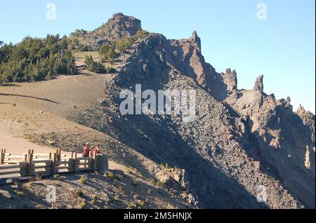 Route pittoresque de l'héritage volcanique - vue sur le backbone du Diable de Watchman. Les gens du Watchman surplombent se tenir derrière un rail de garde et regardent le lac en dessous. Au-dessus d'eux, les dents de lave déchiquetées de la formation de Diable Backbone jab dans le ciel le long du bord du lac Crater. Emplacement : parc national de Crater Lake, Oregon (42,946° N 122,169° O) Banque D'Images