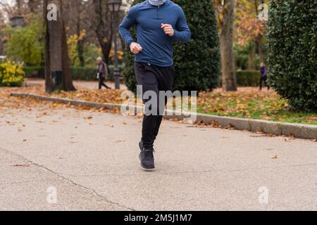 Sportif méconnaissable qui court sur un sentier asphalté recouvert de feuilles sèches lors d'une séance d'entraînement dans un parc d'automne Banque D'Images