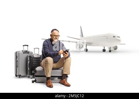 Homme mature assis sur une valise devant un avion et utilisant un smartphone isolé sur fond blanc Banque D'Images