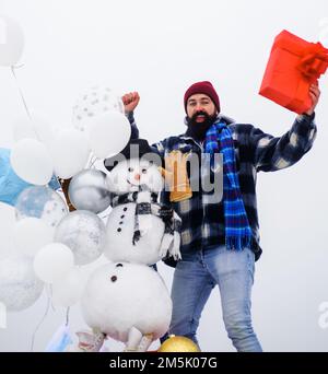 Homme barbu dans des vêtements chauds avec boîte cadeau. Bonhomme de neige en chapeau, écharpe et moufles avec ballons d'air. Banque D'Images