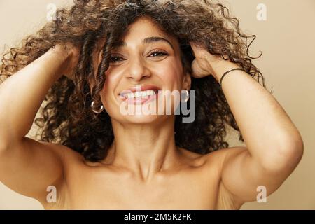 Elle applique de la crème et du baume sur ses cheveux bouclés, le concept de protection et de soin, un look sain, un sourire aux dents sur fond beige Banque D'Images