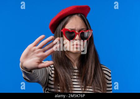 Une femme désintéressée n'approuve pas le geste du signe de la main. Refuser, rejeter, pas d'accord. Portrait d'une jeune femme en lunettes rouges sur fond bleu Banque D'Images