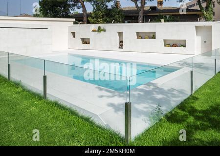 Une piscine en marbre blanc avec une rambarde en verre, un jardin avec des créneaux et des nids de fleurs dans un mur Banque D'Images