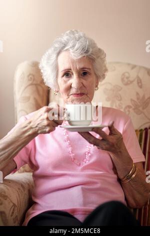 Je fais toujours une bonne tasse de thé. Portrait d'une femme âgée ayant une boisson chaude sur une chaise à la maison. Banque D'Images