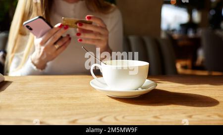 Les mains de la femme tenant le téléphone mobile et la carte de crédit dans le café. Pause café. Tasse de café sur la table. Image floue, mise au point sélective Banque D'Images