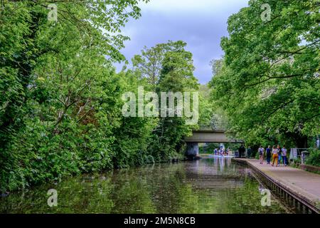 Les gens marchent le long de la piste de remorquage du canal Regent's. Pont du chemin Prince Albert en arrière-plan. Le feuillage de printemps vert et les arbres donnent une atmosphère sereine. Banque D'Images