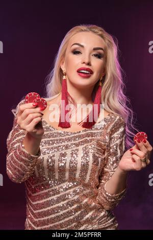 Femme blonde avec une coiffure parfaite et un maquillage lumineux pose avec des jetons de jeu dans ses mains. Casino, poker. Banque D'Images