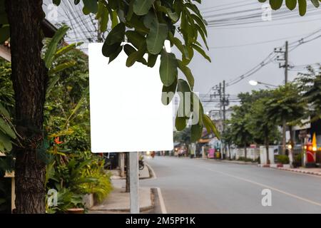 Panneau blanc vierge avec un chemin d'écrêtage derrière les feuilles sur le côté de la rue en ville Banque D'Images