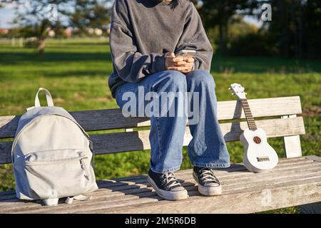 Coupe courte du corps de la jeune fille, assis sur le banc avec ukulele, à l'aide d'un smartphone, mains tenant le téléphone mobile Banque D'Images