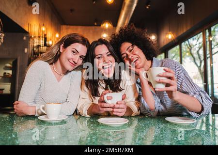 Trois jeunes femmes souriant regardant un appareil photo dans un café. Un groupe de jeunes filles multiraciales heureux s'amusant ensemble à faire une pause dans un restaurant. Photo de haute qualité Banque D'Images