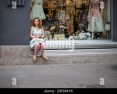 Bucarest, Roumanie - août 2022: REDHEAD Girl assis devant une vitrine de magasin de vêtements dans le centre de Bucarest., Roumanie Banque D'Images