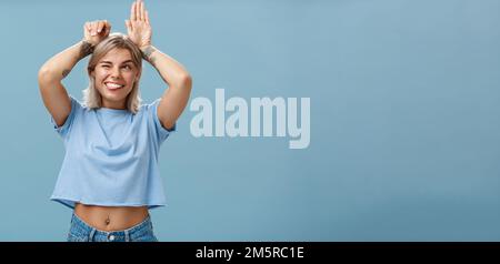 Studio de divertissement insouciant et émouvant femme de charme heureuse avec des tatouages sur les bras agissant comme un lapin avec les paumes sur la tête se fait joies Banque D'Images