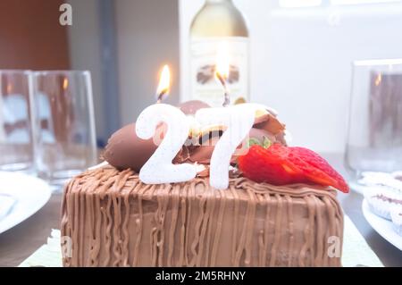 Gâteau d'anniversaire avec bougies en forme de numéro 27 sur une table montée pour quelques personnes et une bouteille de vin en arrière-plan, concept d'anniversaire pour adultes Banque D'Images