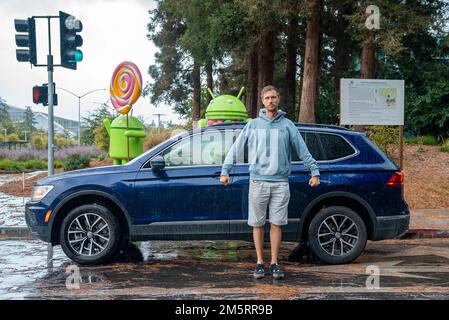 Homme devant une voiture bleue sur une rue mouillée avec des statues android en arrière-plan Banque D'Images