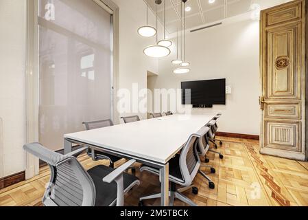 Salle de réunion d'un imposant bâtiment d'époque avec une longue table en bois blanc avec chaises pivotantes grises, parquet et lampes design Banque D'Images