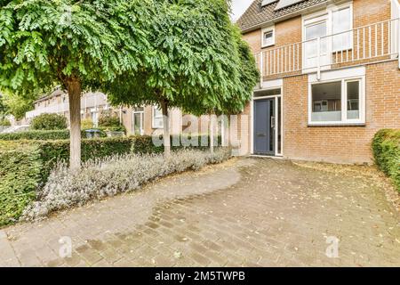 une vue extérieure d'une maison avec des arbres et des buissons dans la cour avant, sur un chemin pavé de briques menant à une porte bleue Banque D'Images