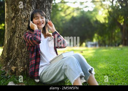 Bonne femme asiatique Profitez de la musique dans un casque tout en étant assise sous l'arbre sur l'herbe verte à une belle journée ensoleillée Banque D'Images
