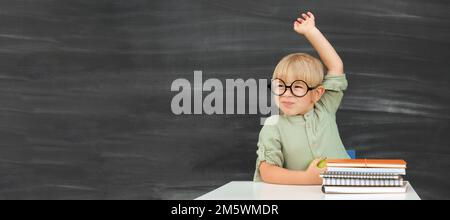 Retour à l'école. Joyeux élève souriant dans des verres ronds assis au bureau. L'enfant lève la main dans la salle de classe avec le tableau noir sur fond. Enfant Banque D'Images